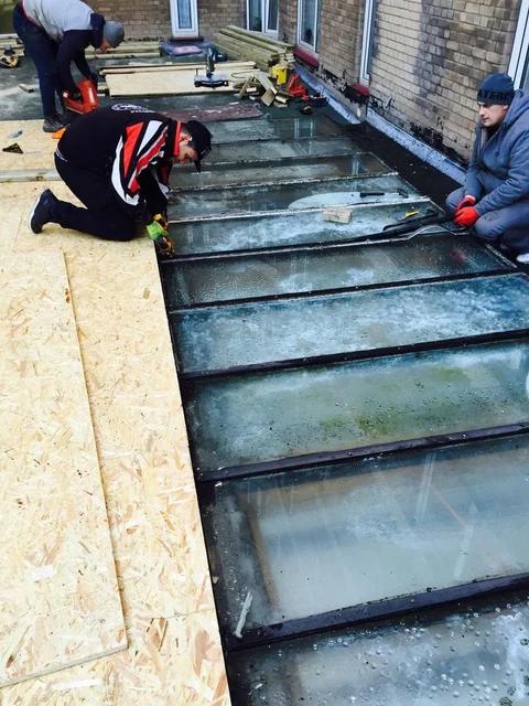 Workers fitting boards on a roof beside a glazed roof opening.