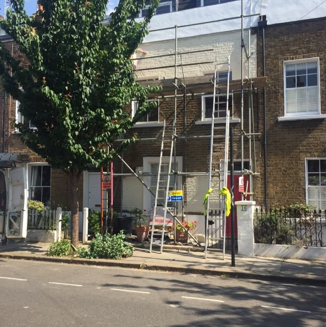 Scaffolding against a brick building with a pale upper wall and a tree at the left