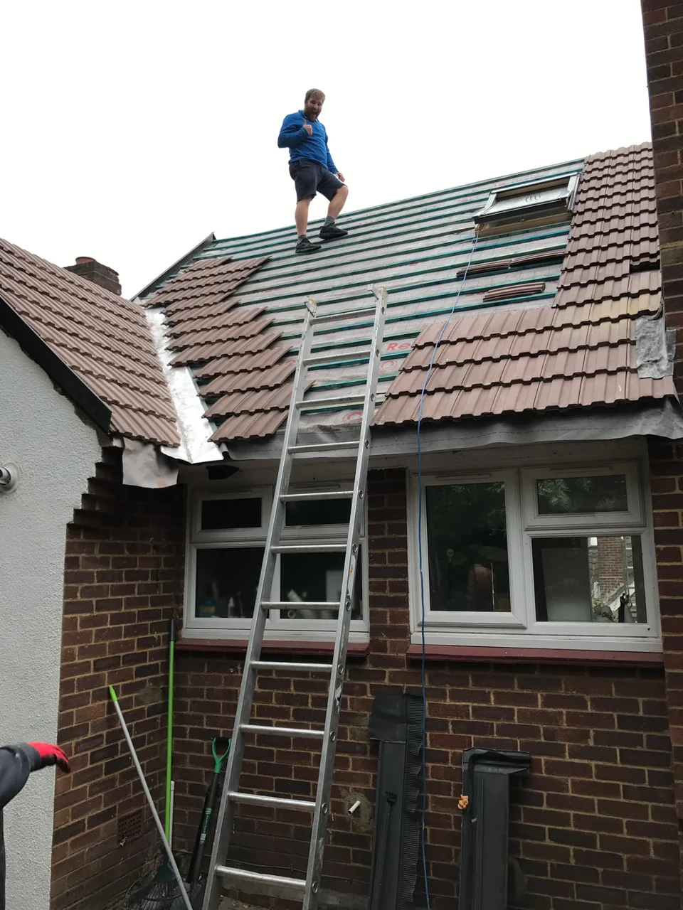 Worker on a partially stripped pitched roof beside a ladder and replacement tiles.