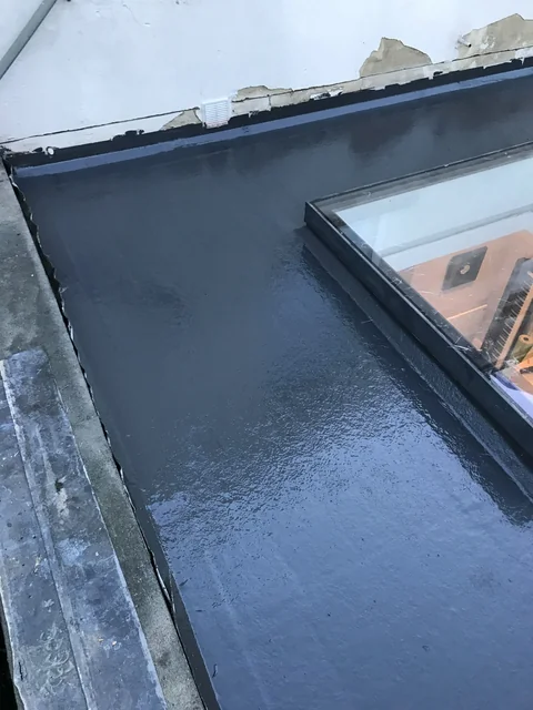 Dark flat-roof surface around a glazed rooflight beside a white wall with exposed patches